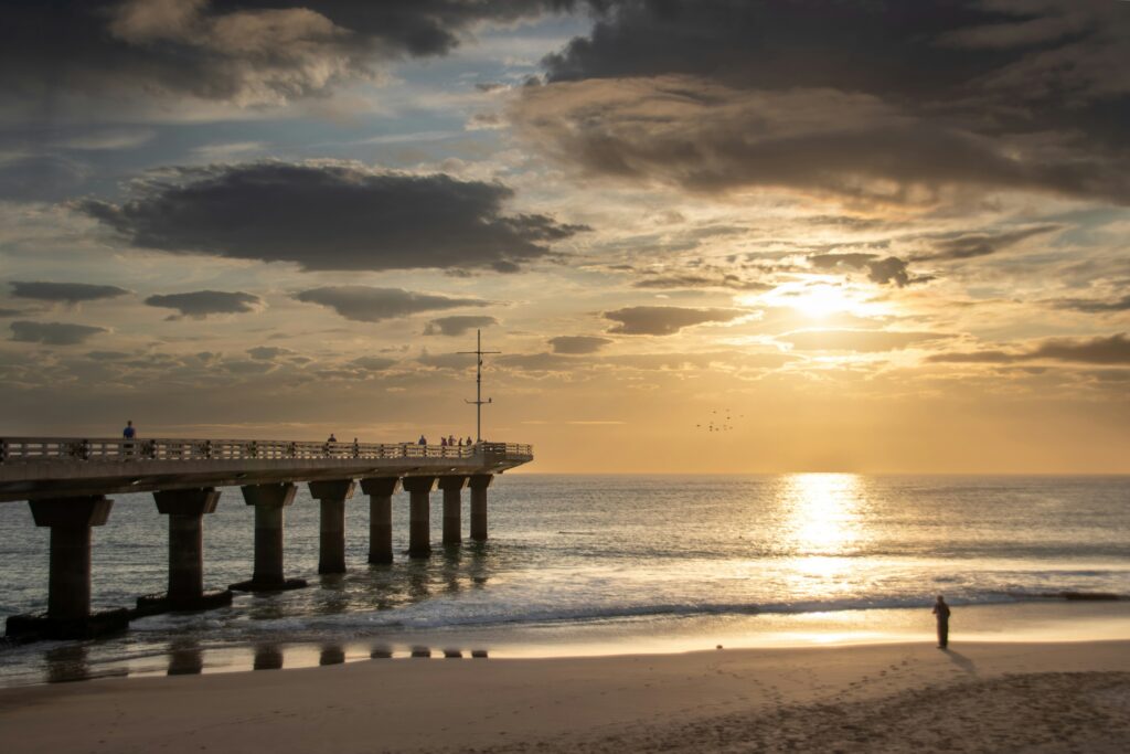 Shark Rock Pier na Hobie Beach, em Gqeberha, ao nascer do sol, com pessoas no píer e a luz refletida na Baía de Algoa.