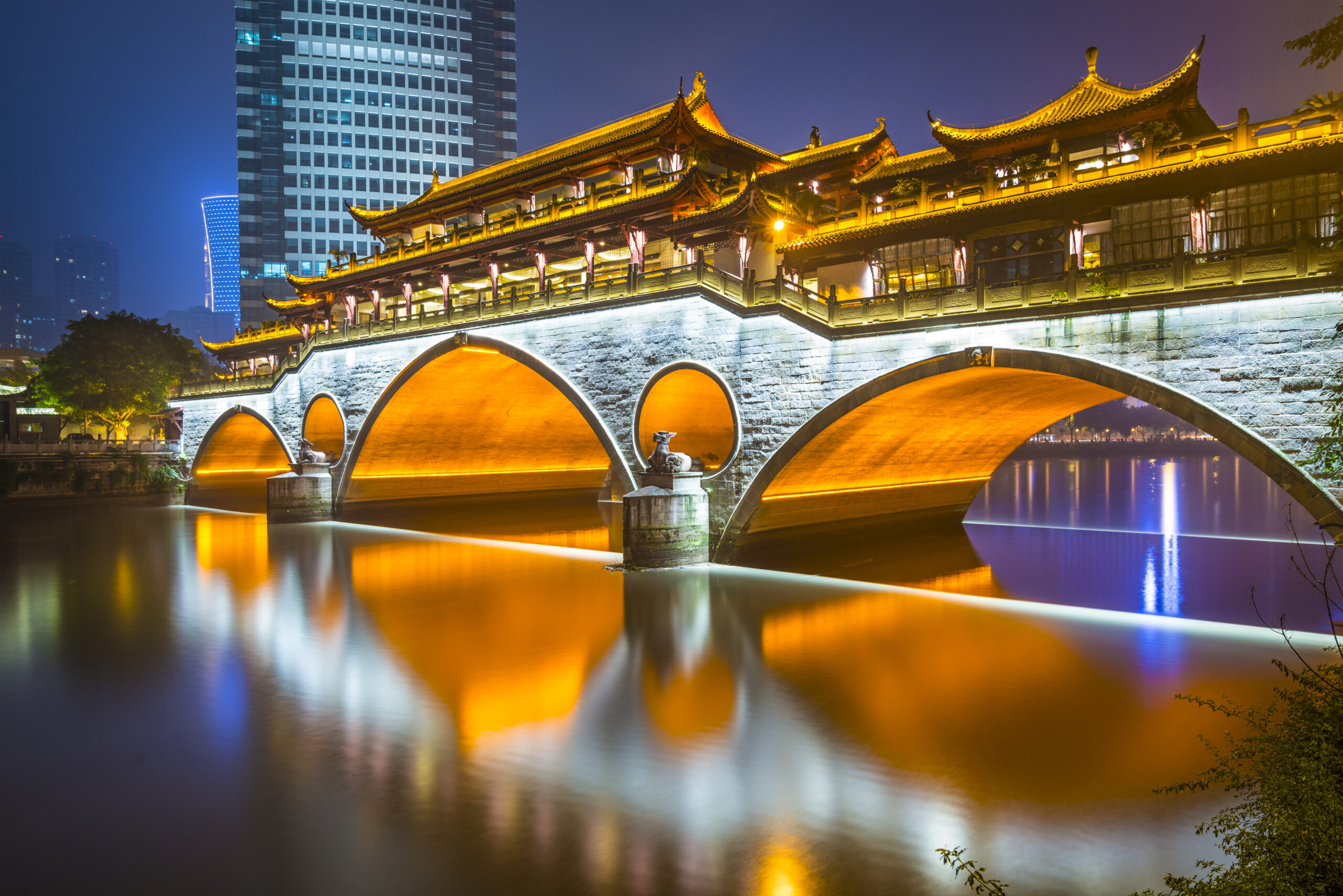 Ponte Anshun iluminada à noite sobre o Rio Jin, em Chengdu, com pavilhões tradicionais e arranha-céus ao fundo.