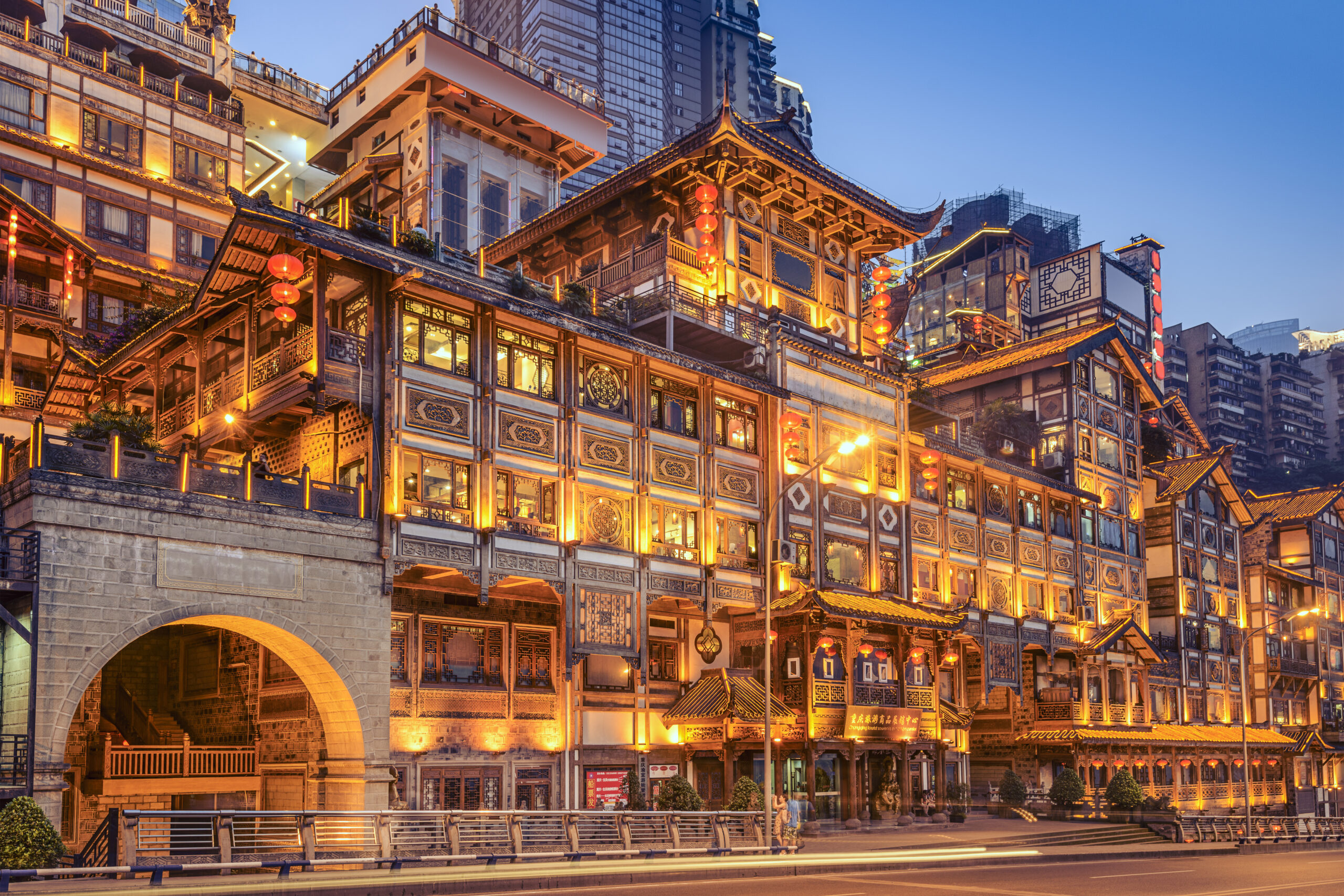 Fachada de Hongyadong iluminada ao anoitecer, com construções tradicionais de madeira, lanternas vermelhas e prédios modernos ao fundo, em Chongqing.