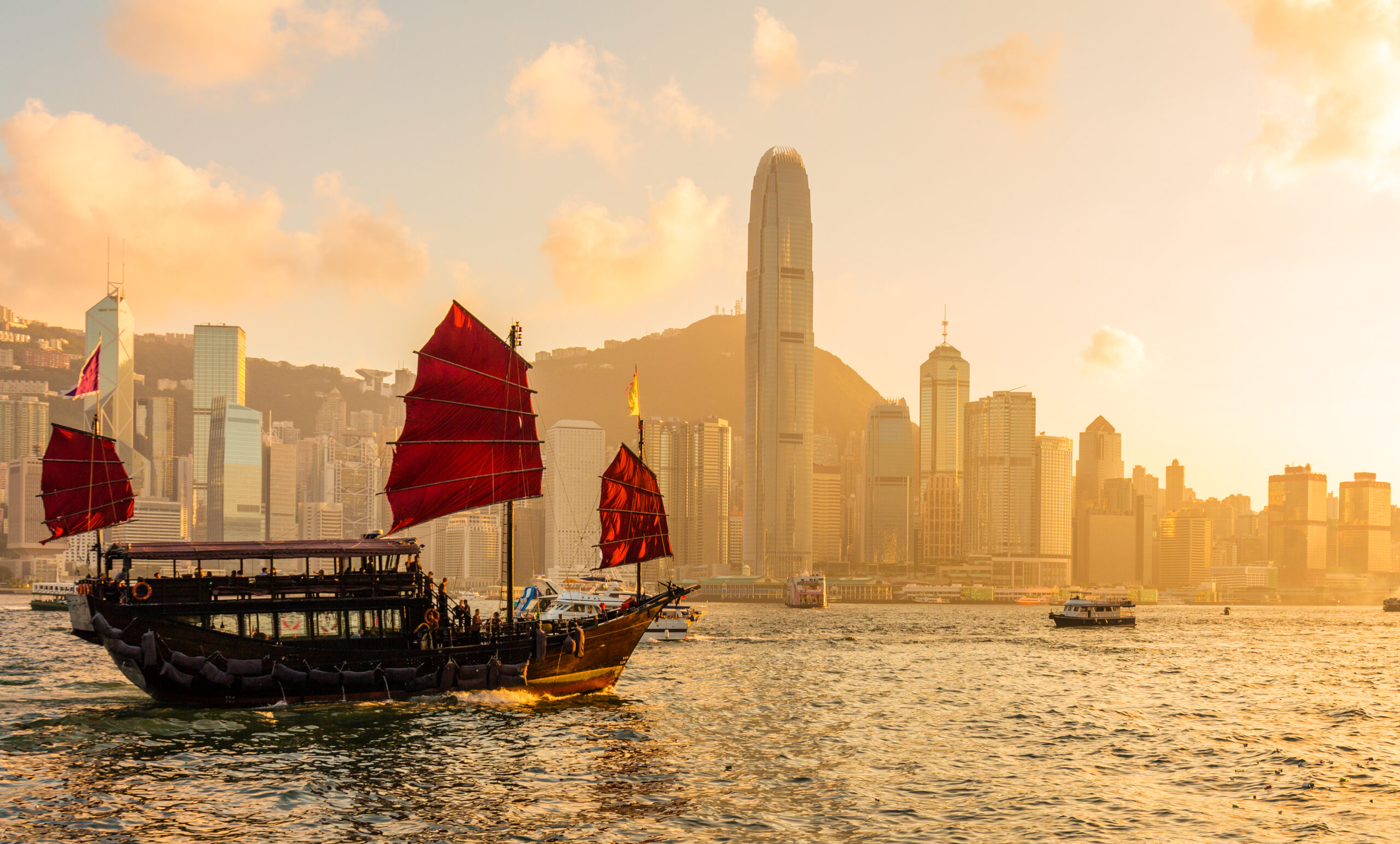 Junco de velas vermelhas navegando no Porto Victoria ao pôr do sol, com os arranha-céus da Ilha de Hong Kong ao fundo.
