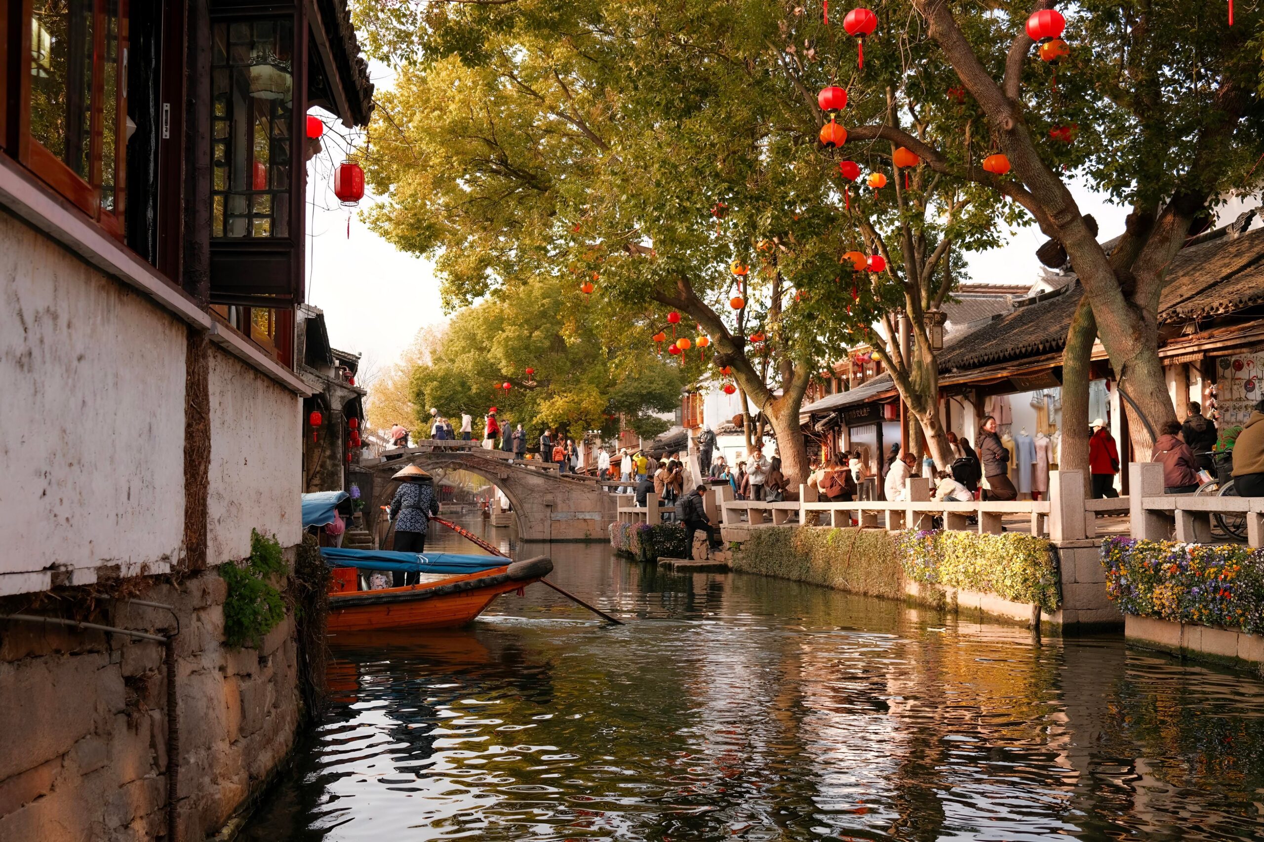 Barqueiro conduzindo um barco por um canal de Suzhou, com ponte de pedra em arco, lanternas vermelhas nas árvores e casas tradicionais às margens.
