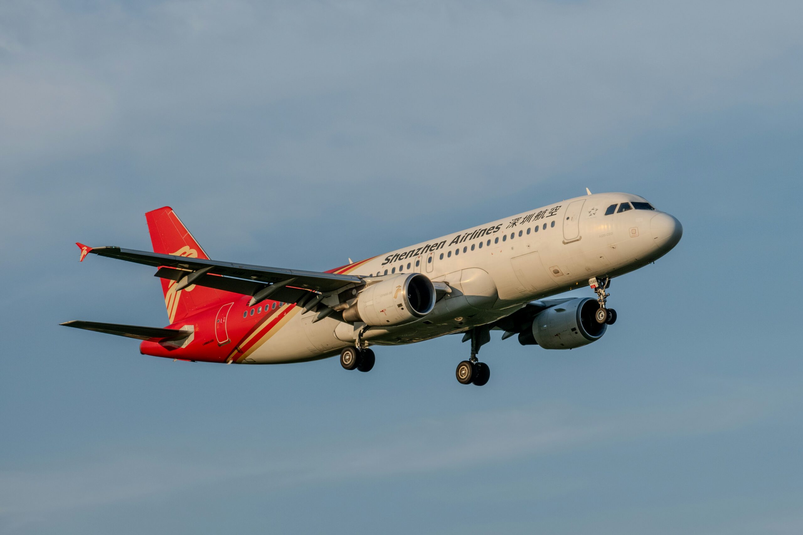 Avião da Shenzhen Airlines em voo, com o trem de pouso abaixado, contra o céu azul.