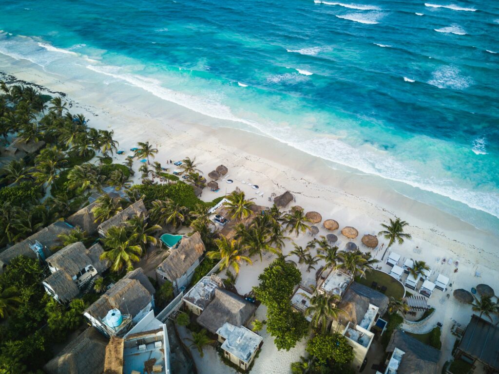 Vista aérea de hotéis boutique com palapas entre coqueiros na praia de Tulum, com mar turquesa do Caribe mexicano