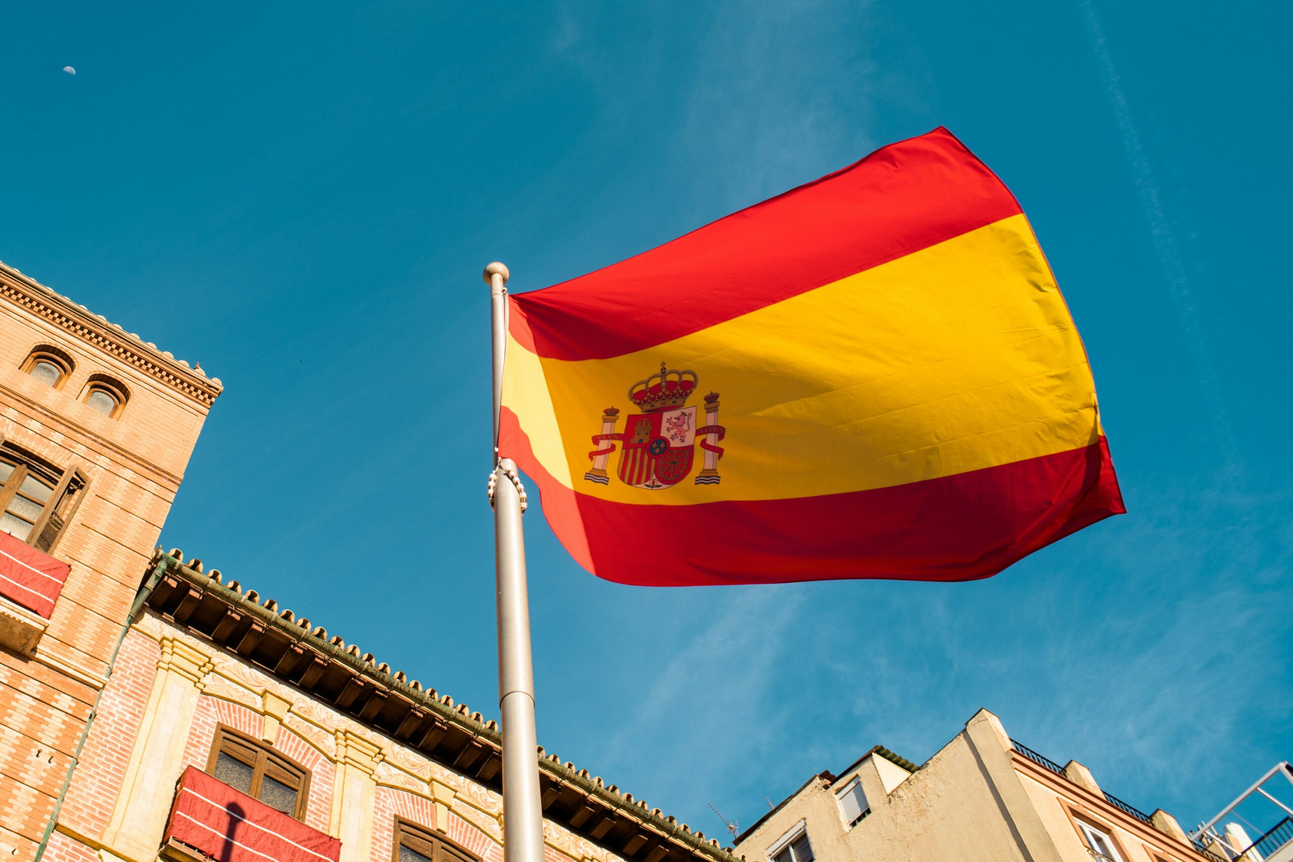Bandeira da Espanha hasteada em frente a edifício histórico com céu azul ao fundo