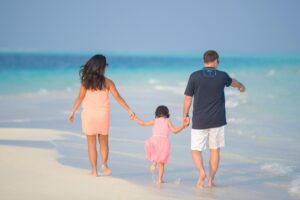 Família com criança pequena caminhando de mãos dadas na beira do mar em praia de areia branca e água azul-turquesa.