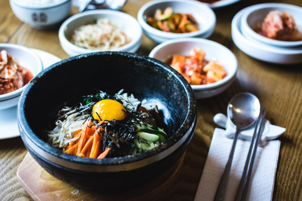 Bibimbap servido em tigela de pedra quente com gema, legumes e alga, cercado por banchan como kimchi e kimchi de rabanete.