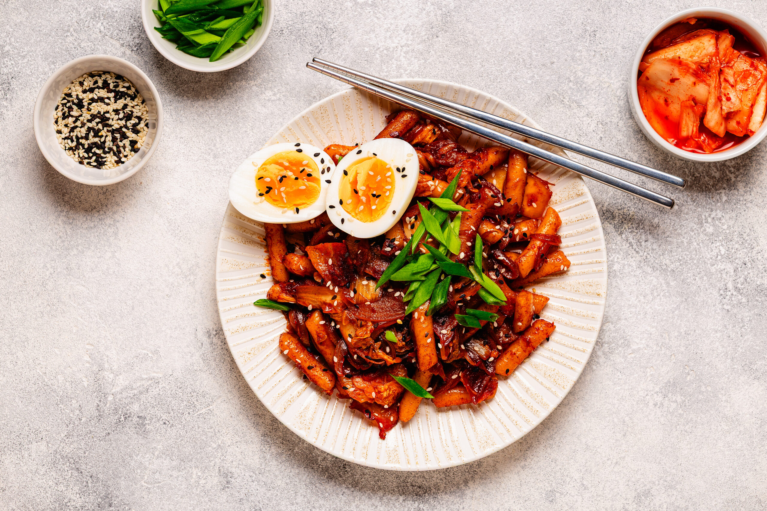 Tteokbokki, bolinhos de arroz em molho picante, com kimchi, ovo cozido, cebolinha e gergelim, ao lado de uma tigela de kimchi.